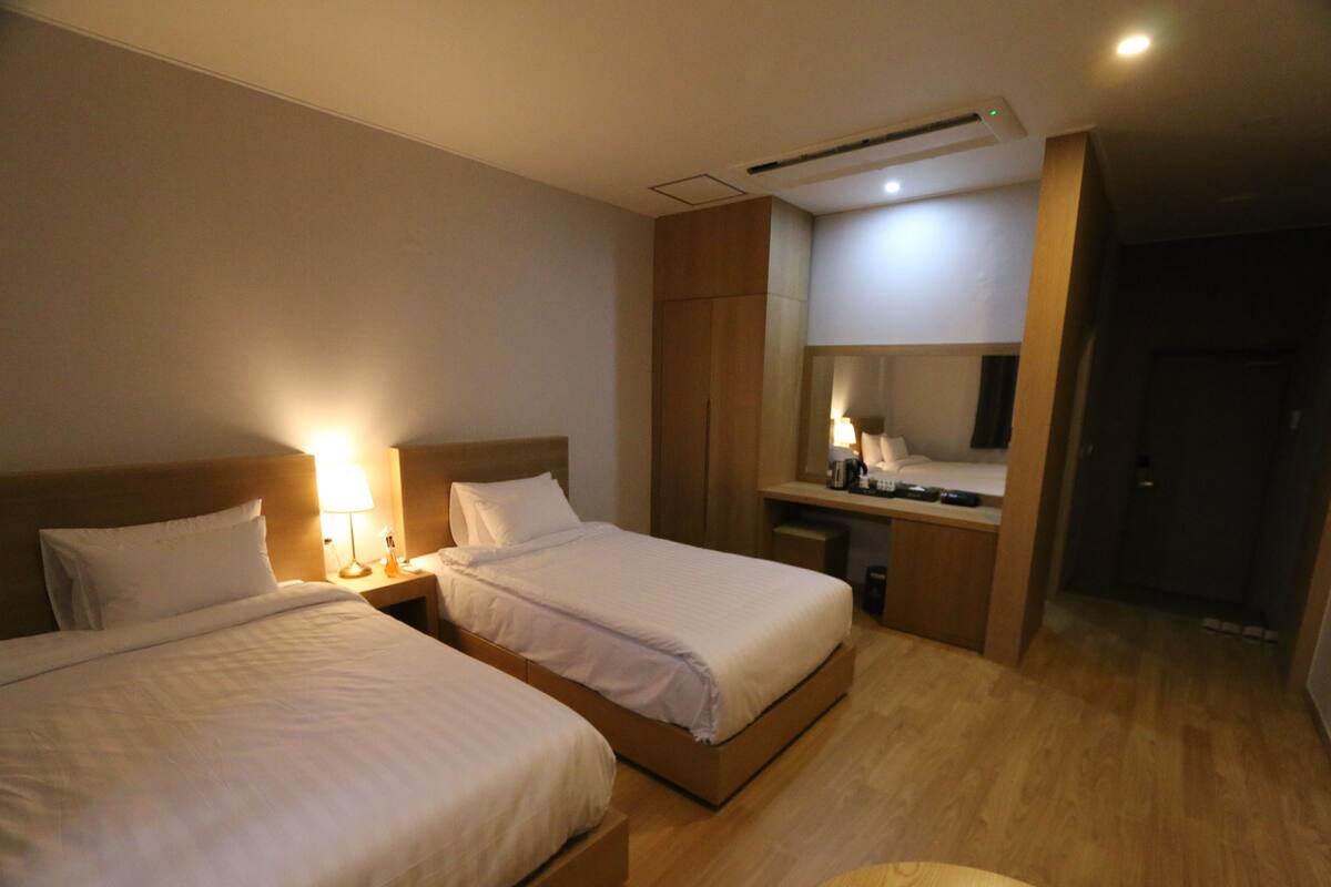 A hotel room with two beds, a desk, a mirror, and a small kitchenette.