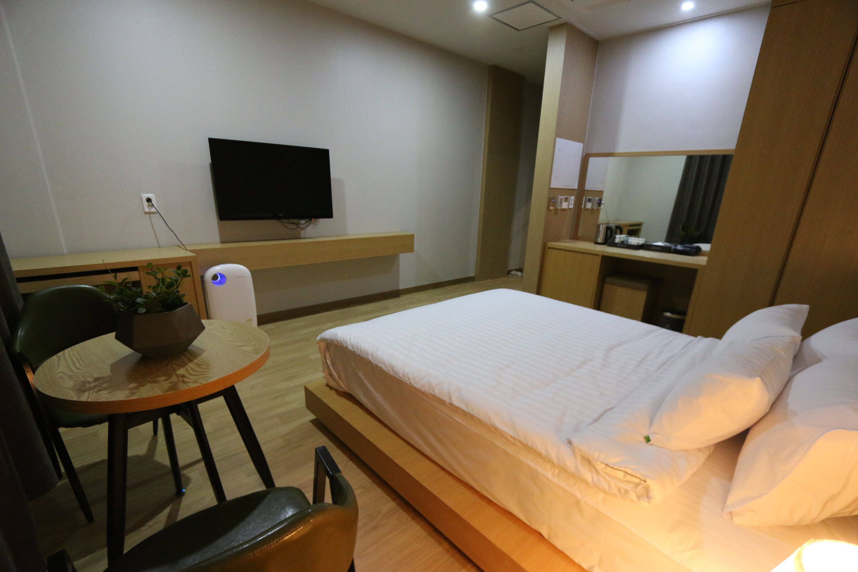 A hotel room with a bed, a desk with a computer, a chair, a small table with a plant, and a TV mounted on the wall.