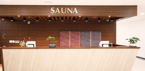Un mostrador de recepción de madera con un letrero que dice "SAUNA" encima. Hay plantas en macetas sobre el mostrador, un estante montado en la pared con botellas y una pequeña señal con un símbolo rojo.