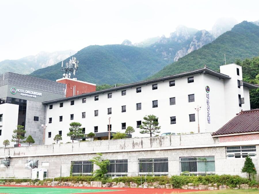 A hotel with a modern design, featuring a flat roof and multiple windows. It is situated in a mountainous area with lush greenery.