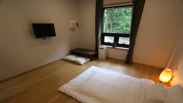 Un dormitorio con cama, televisión de pantalla plana en la pared, ventana con vista a árboles y lámpara de noche.