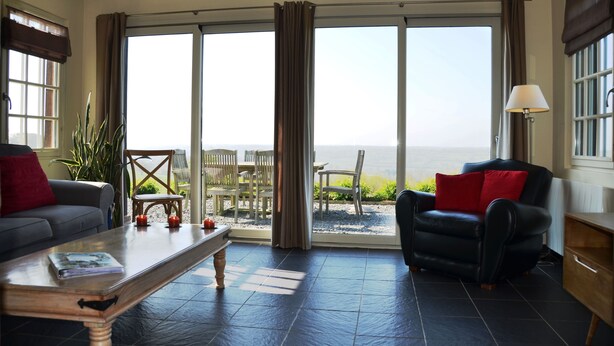 A living room with a sofa, armchair, and coffee table, large sliding glass doors leading to a patio with outdoor furniture.