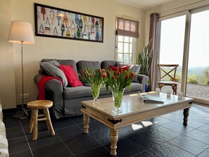 Ein Wohnzimmer mit grauem Sofa, roten Kissen, einem Holztisch, einem Strauß Tulpen, einer Stehlampe und einem Fenster mit Blick auf Grünflächen.