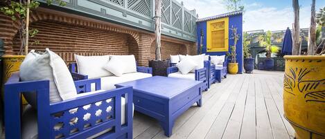 Un área de estar al aire libre con mobiliario azul y cojines blancos, una terraza de madera y un edificio con detalles azules y amarillos al fondo.