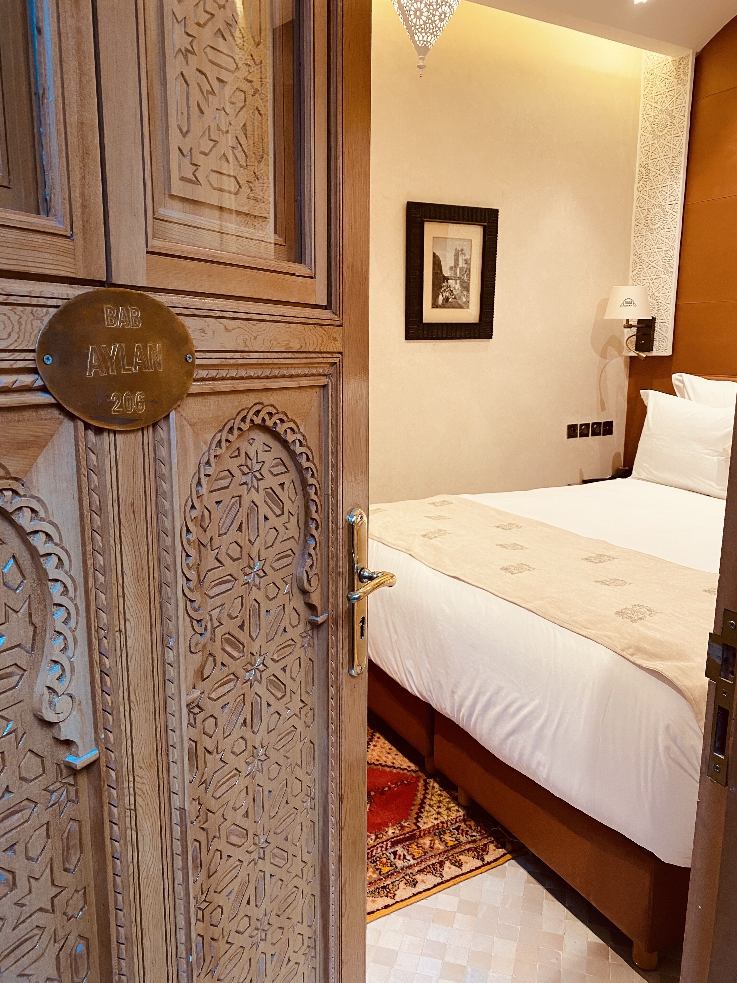 A hotel room with a wooden door, a bed with white bedding, a patterned rug, and a framed picture on the wall.