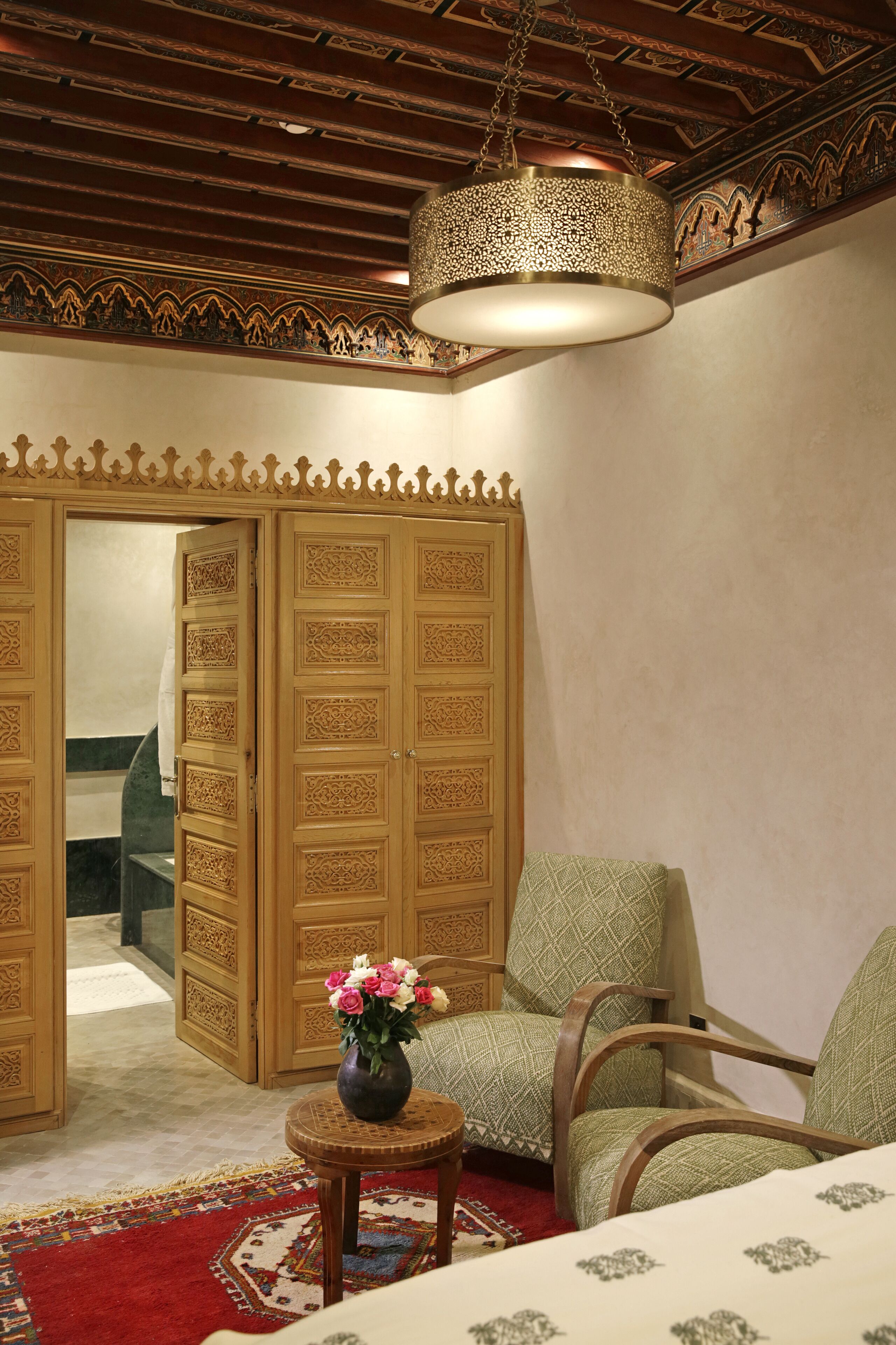 A room with a patterned wooden wardrobe, a green upholstered chair, a small round table with a vase of flowers, and a hanging light fixture.