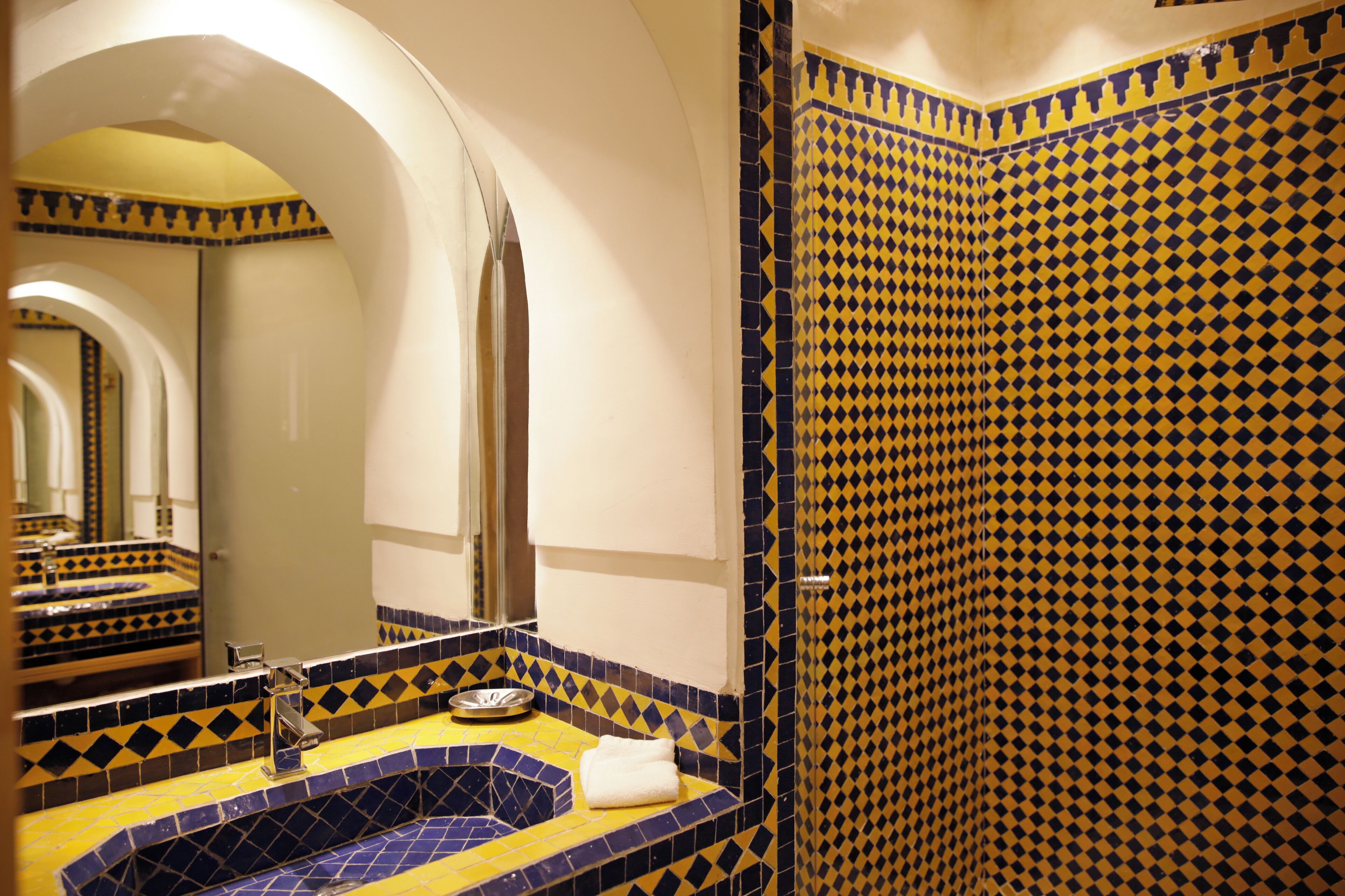 Ein Badezimmer mit einem Becken aus gelb-blauen Fliesen, einem Spiegel und einer Dusche mit Rautenmuster.