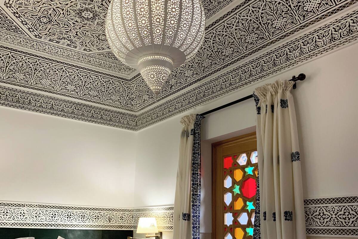 Une pièce dotée de motifs complexes sur le plafond, d’un grand lit recouvert de draps blancs, d’un luminaire suspendu et d’une fenêtre ornée de vitraux colorés.