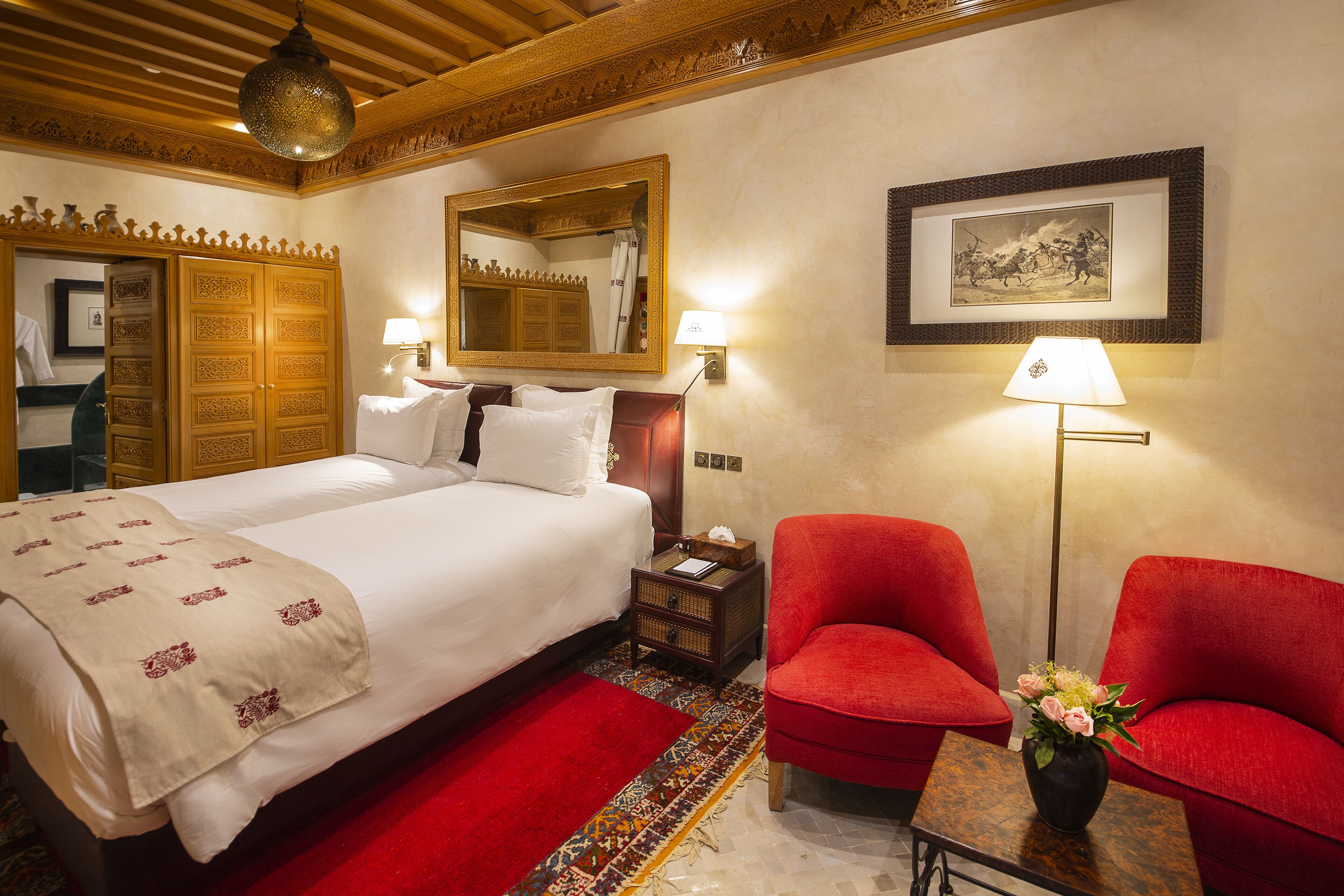A hotel room with a large bed, two red armchairs, a small table with a vase of flowers, a nightstand, a mirror, and a framed picture on the wall.