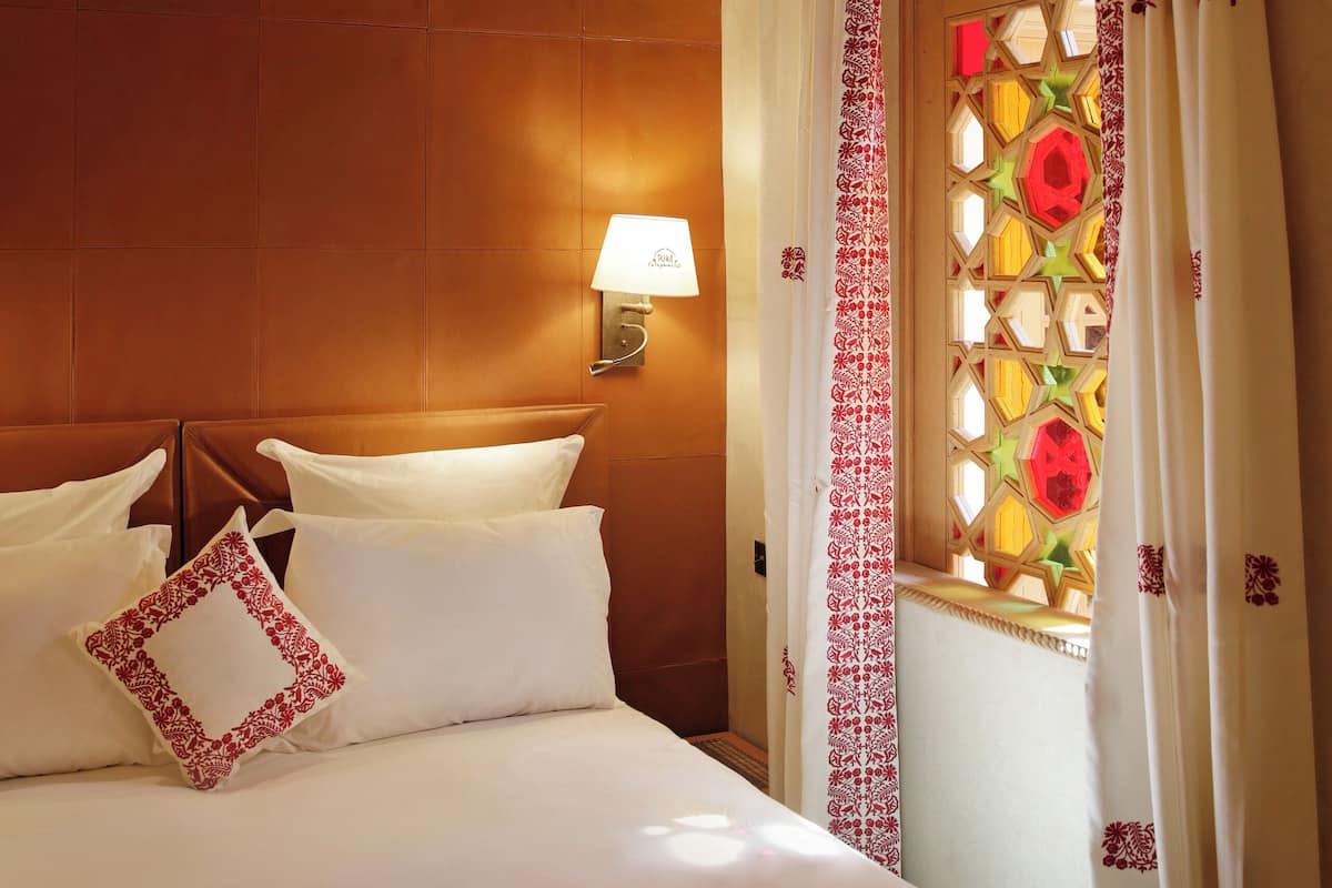 Une chambre d’hôtel avec un lit, une lampe fixée au mur et une fenêtre agrémentée de rideaux décoratifs.