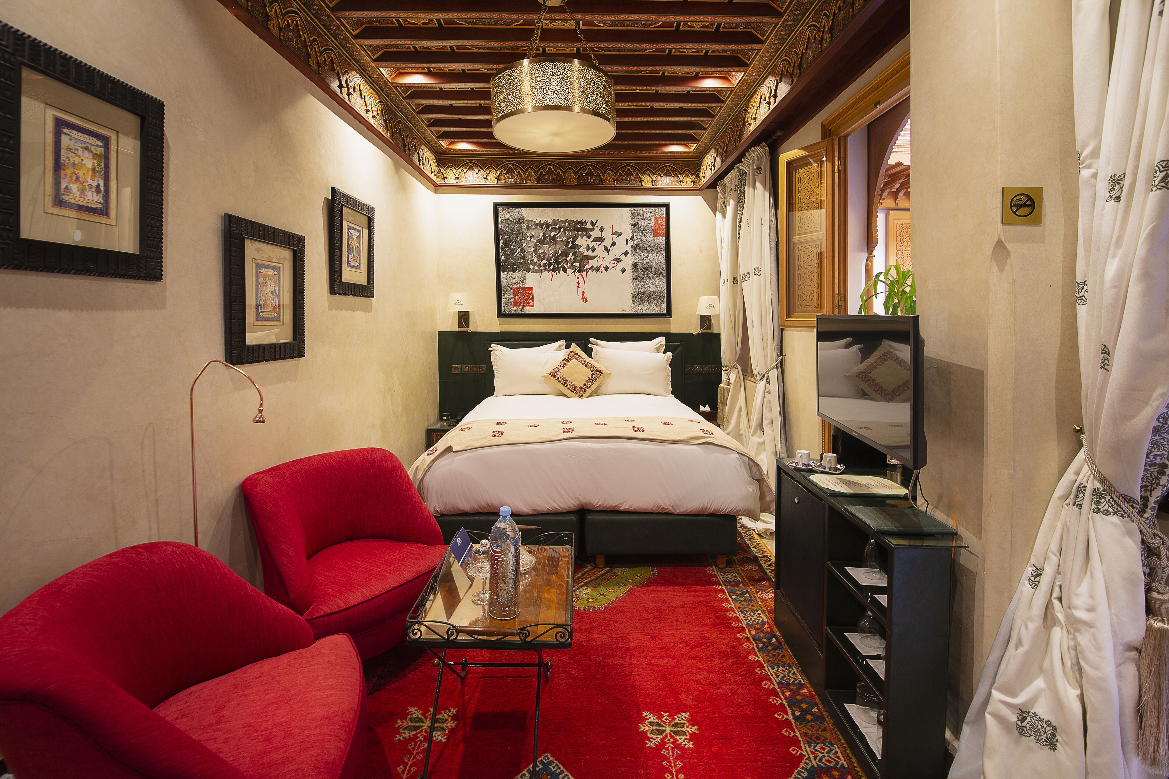 A hotel room with a bed, red chairs, a small table with a bottle, a TV, and artwork on the walls.