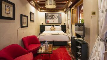 A hotel room with a bed, red chairs, a small table with a bottle, a TV, and artwork on the walls.