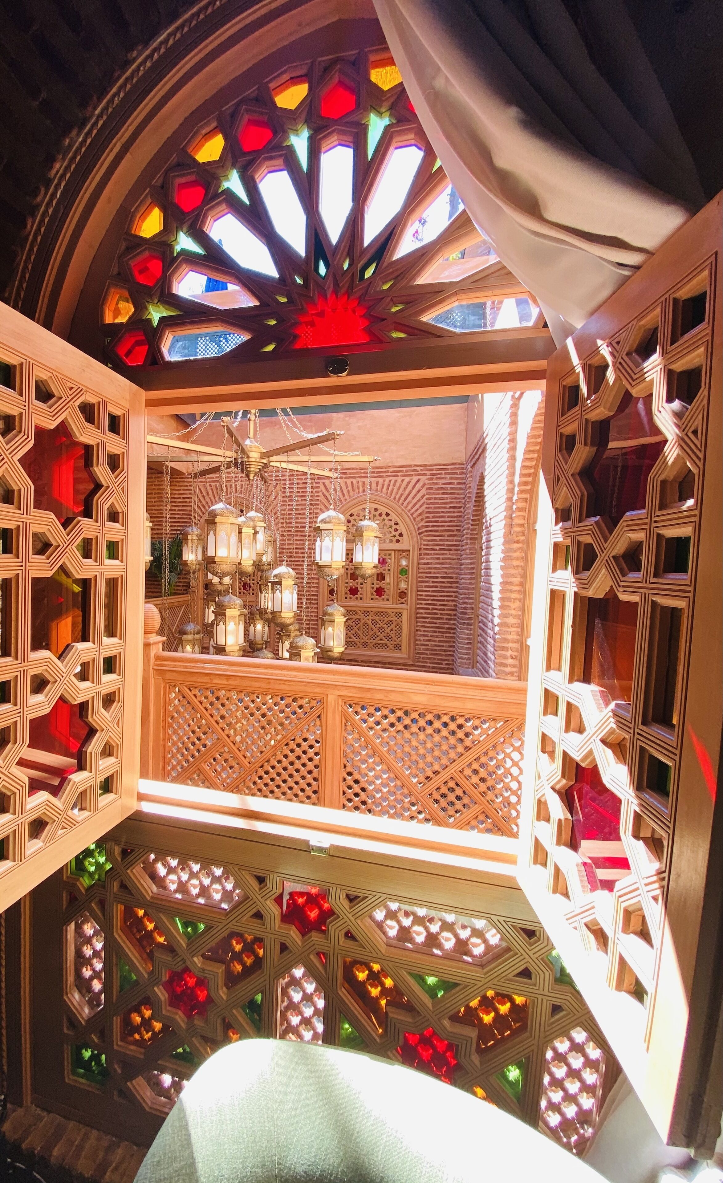 A room with intricate wooden lattice work, a hanging lantern chandelier, and a view through a geometric window with colorful glass panes.