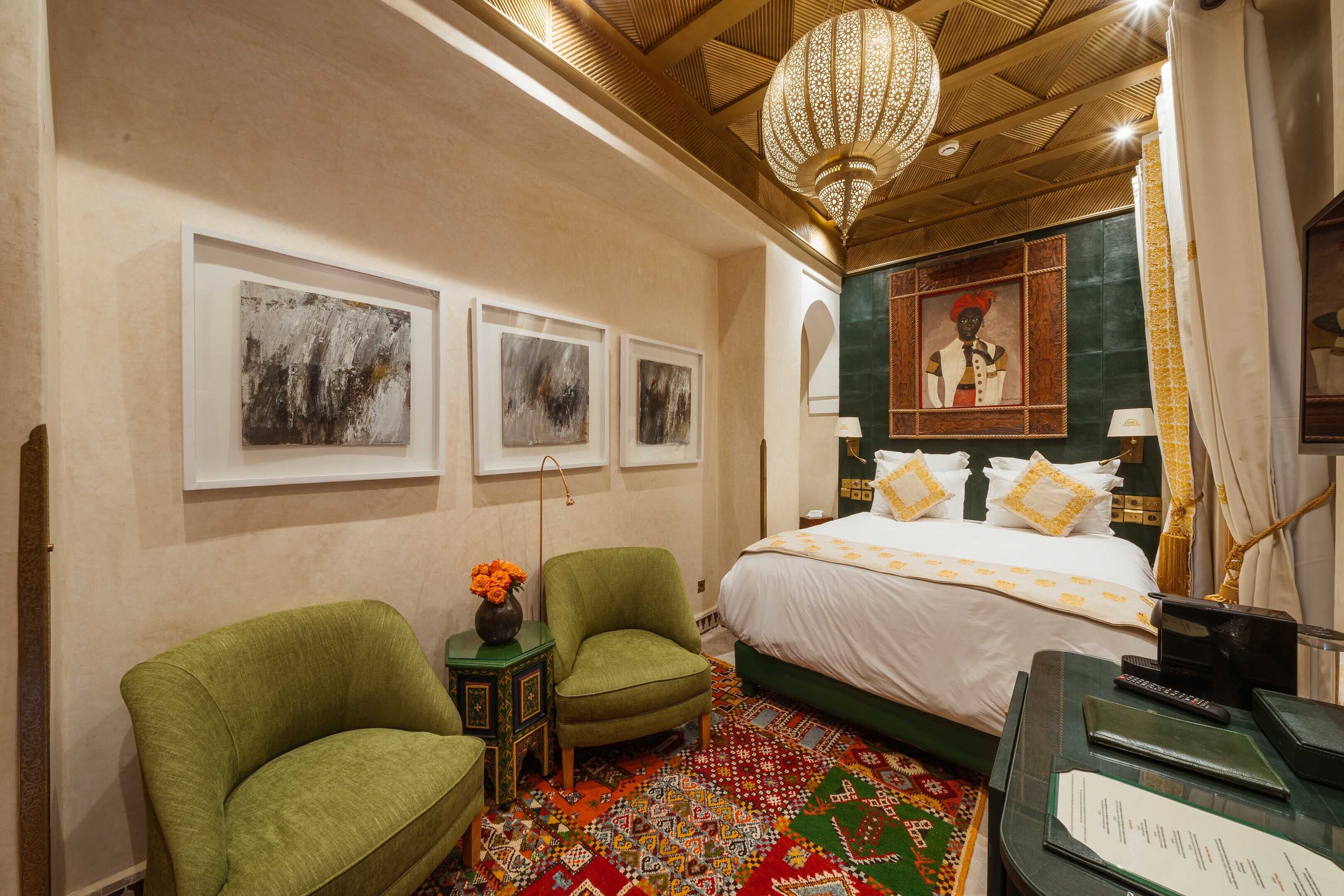 Habitación de hotel con una cama, dos sillones verdes, una alfombra estampada y obras de arte en las paredes.