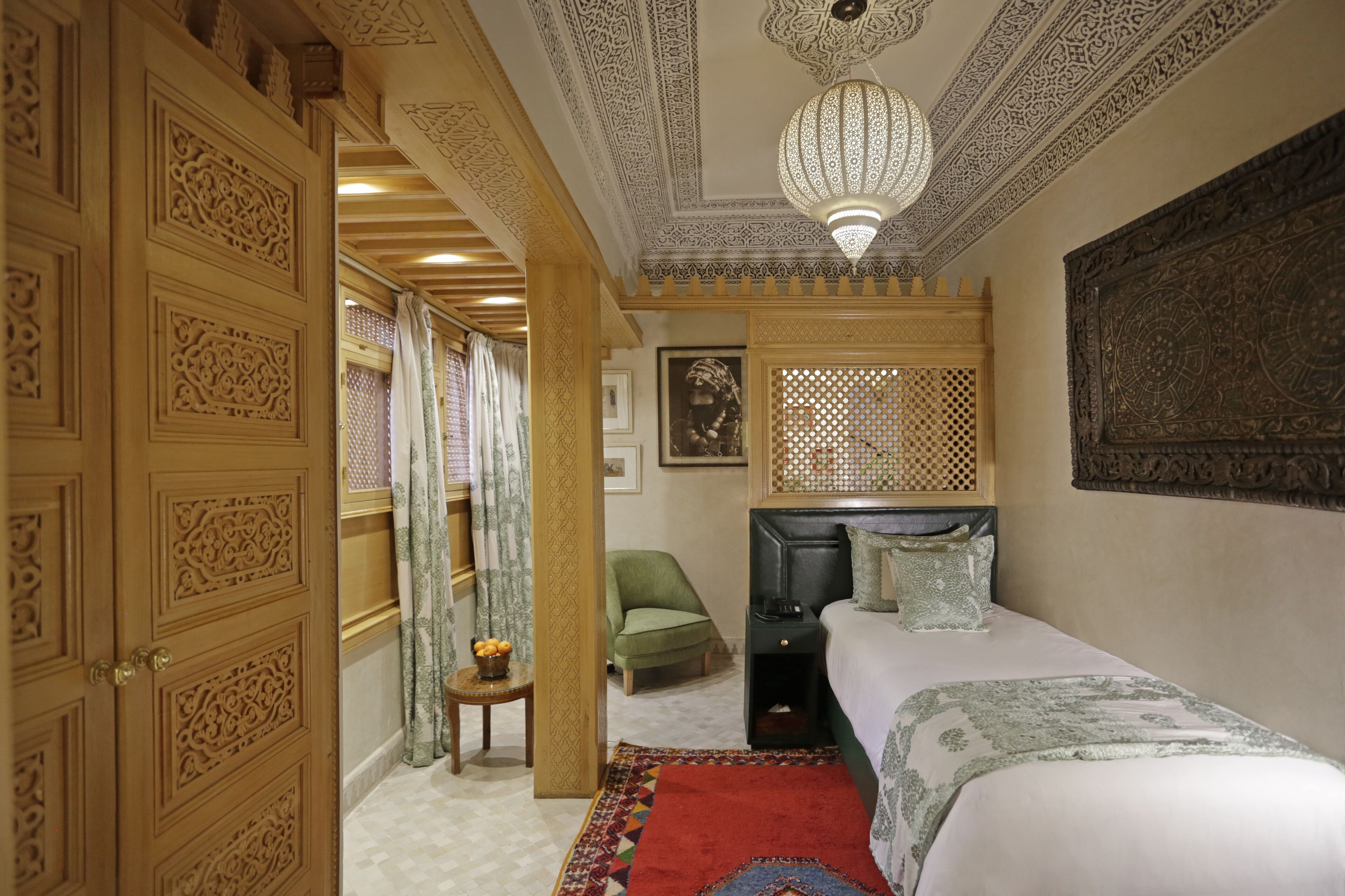 A room with ornate wooden doors, a bed with a patterned bedspread, a green armchair, and a decorative ceiling light.