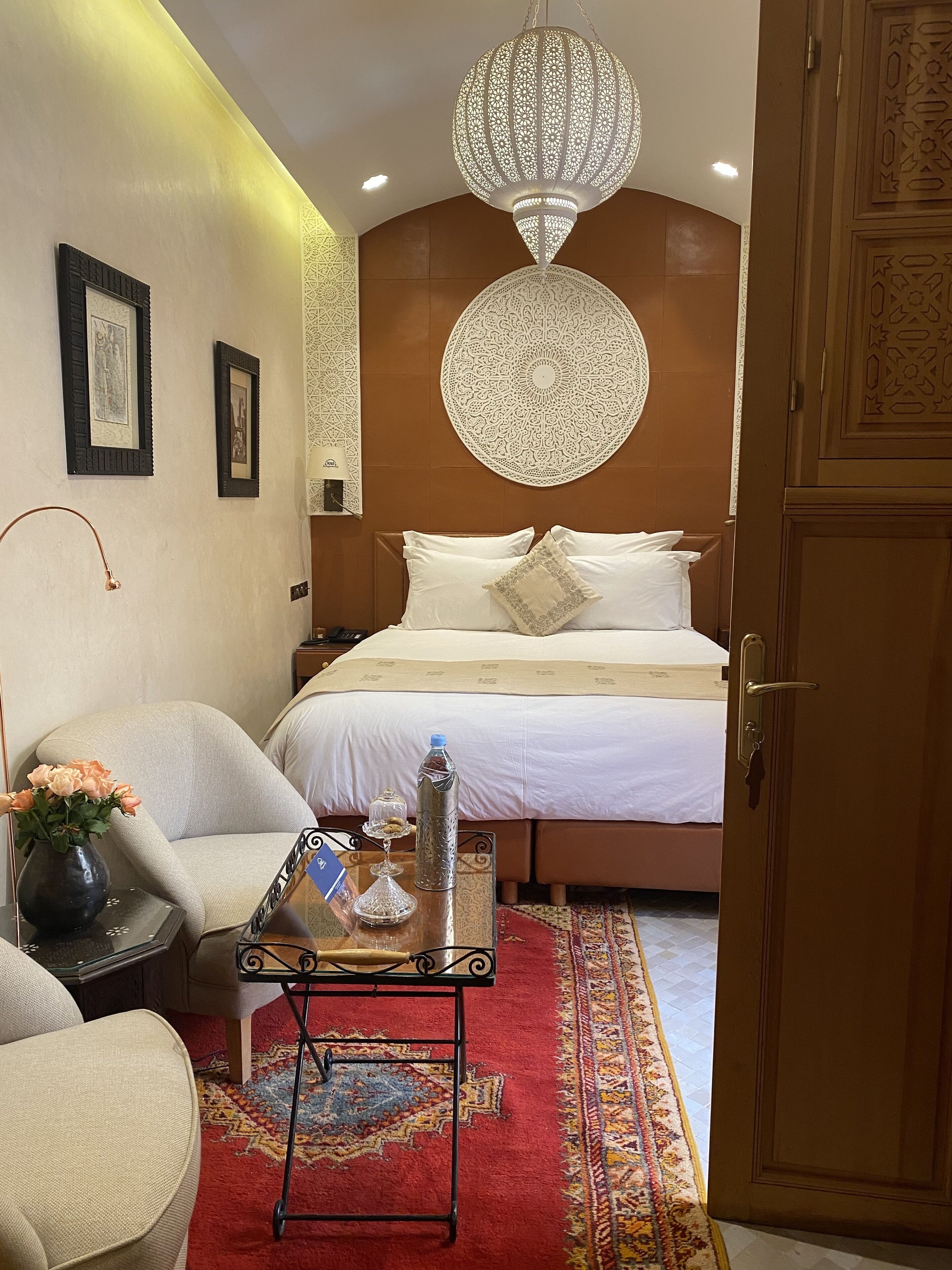 A hotel room with a bed, a nightstand, a chair, a table with a vase of flowers, a water bottle, and a decorative wall hanging.