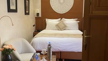 A hotel room with a bed, a nightstand, a chair, a table with a vase of flowers, a water bottle, and a decorative wall hanging.