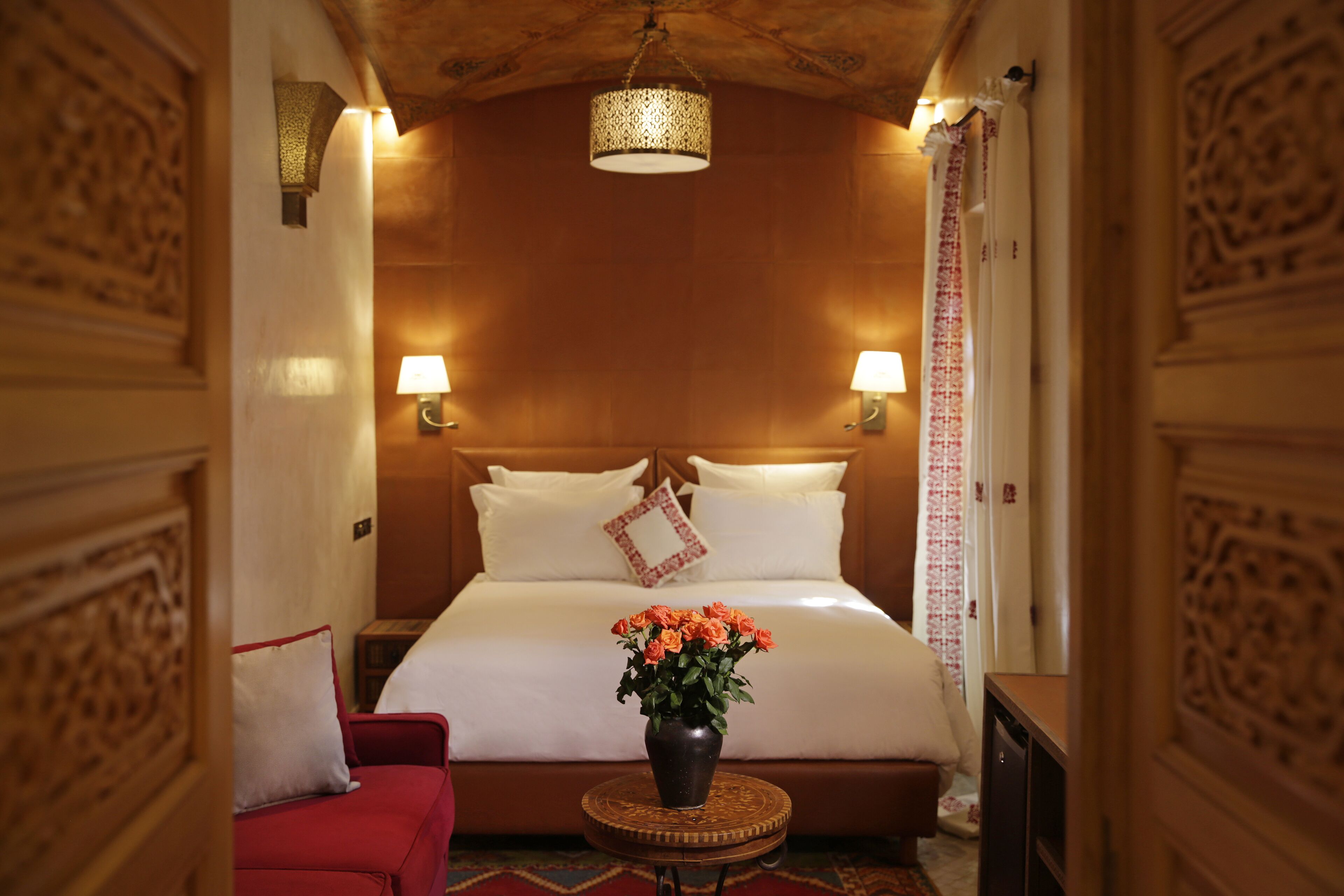 Una habitación de hotel con una cama grande, dos lámparas de noche, un florero con flores, un sofá rojo y un cabecero de madera.
