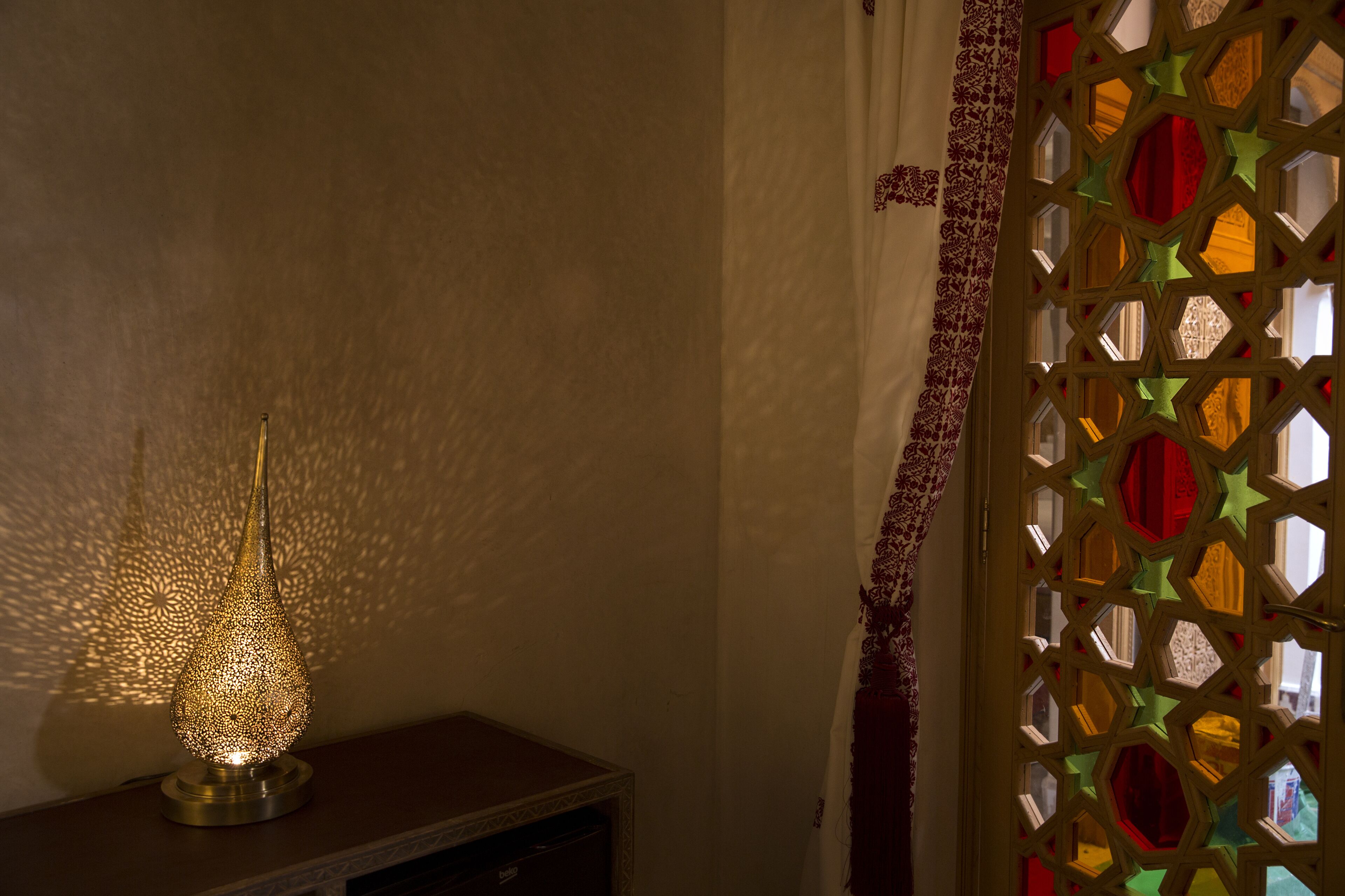 Een decoratieve lamp op een plank, een behang met patroon en een raam met geometrisch ontwerp.