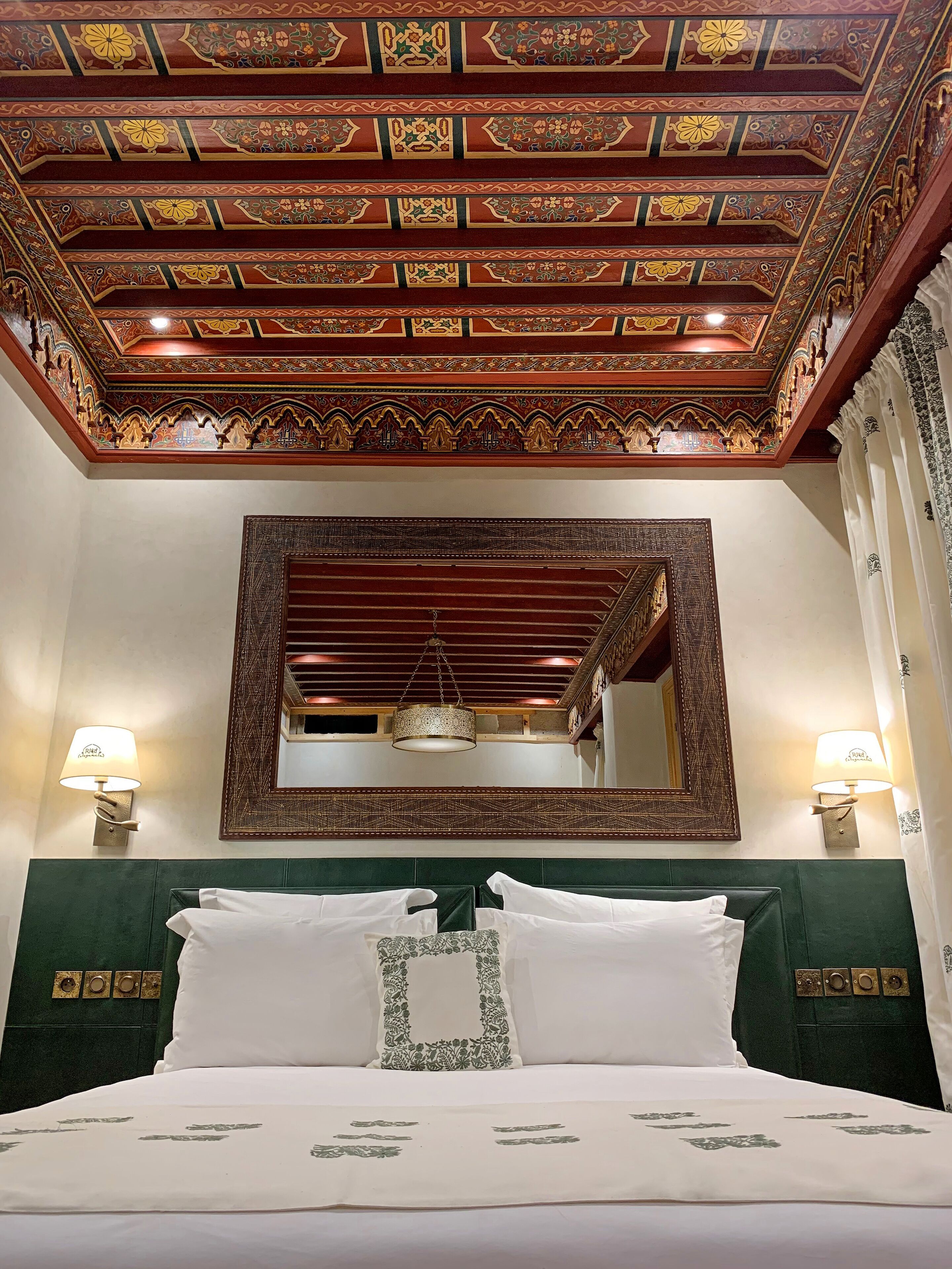 A hotel room with a large bed, green headboard, ornate ceiling, and a large mirror.