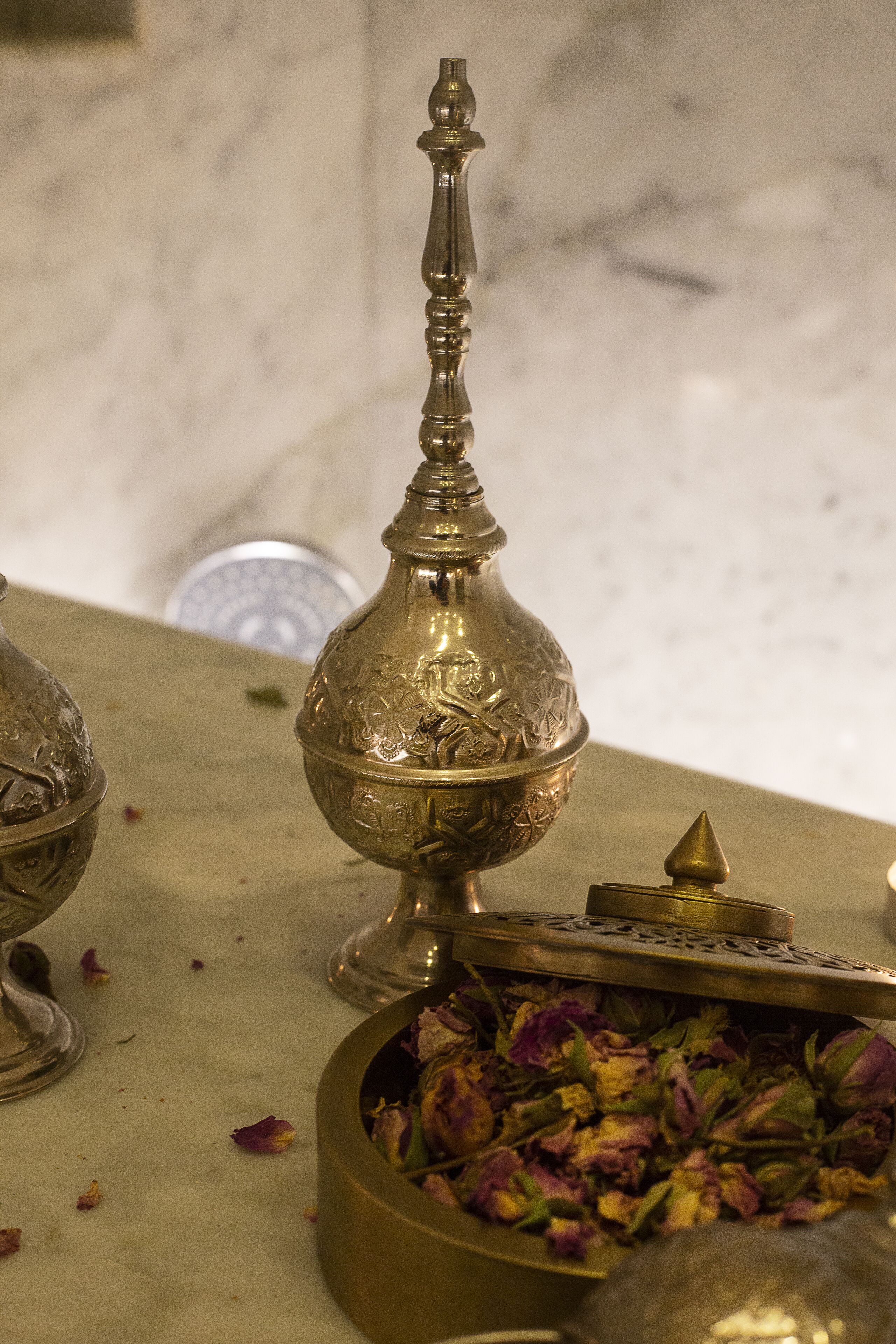 A brass container with intricate designs, a small bowl with dried flowers, and a decorative metal piece on a marble surface.