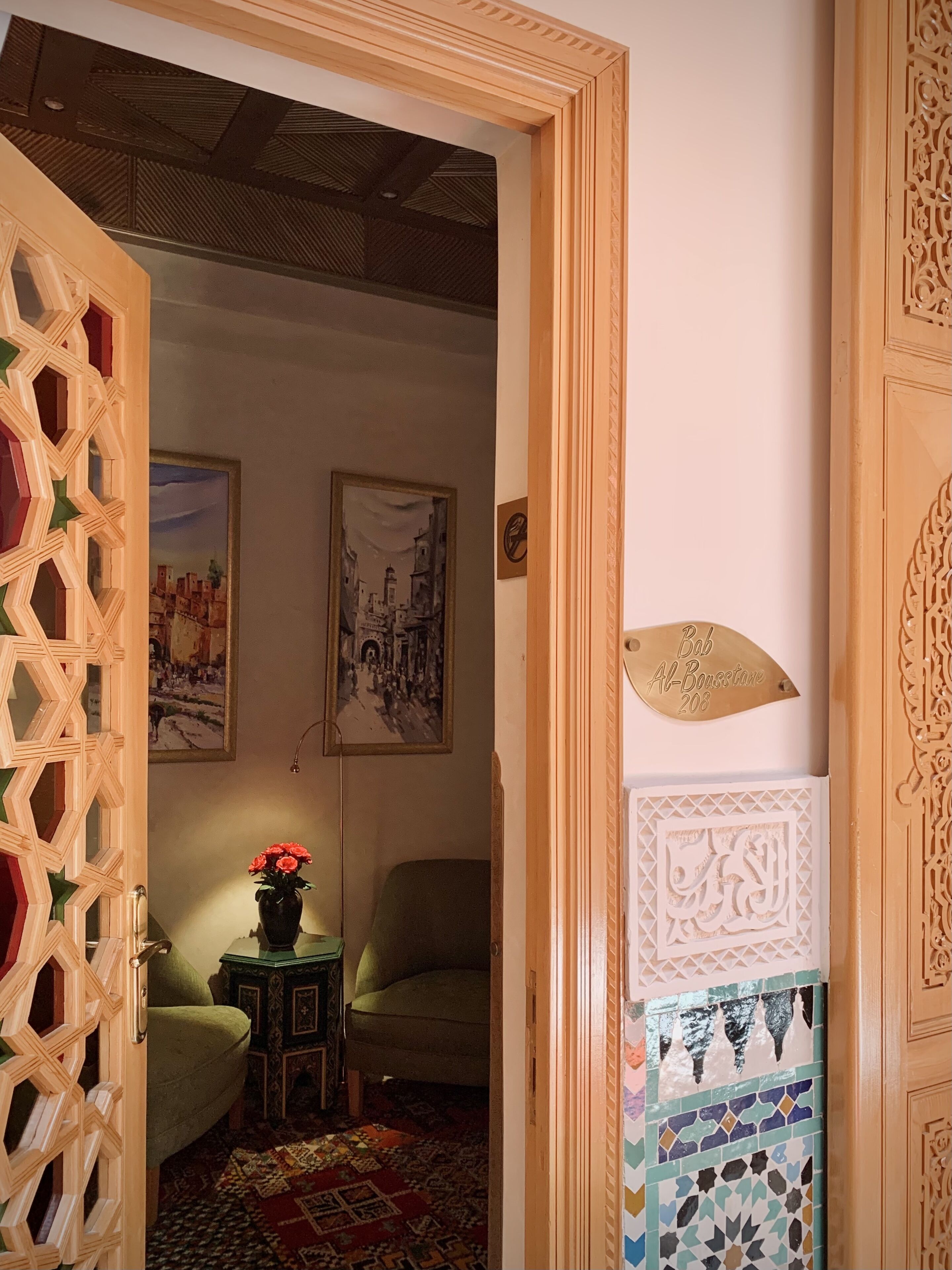 Una habitación con una puerta con diseño, un sillón verde, una mesita con un florero y una placa en la pared.