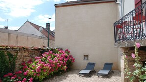 Un patio con dos sillones de descanso, un muro de piedra con plantas en flor y un edificio con balcón rojo.