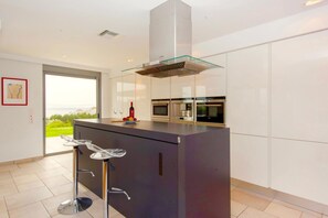 A modern kitchen with a central island, built-in ovens, and a glass-fronted range hood. - Villa Stella (Chania)
