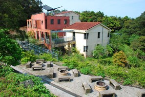 A building with a red and white facade, a balcony, and a fire pit area with seating. - YHA Sze Lok Yuen Tai Mo Shan Youth Hostel (Tsuen Wan)