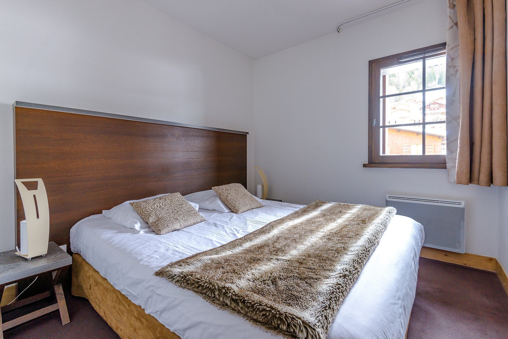 A bedroom with a wooden headboard, a large bed with white linens, a fur throw, a bedside table with a lamp, and a window with curtains.