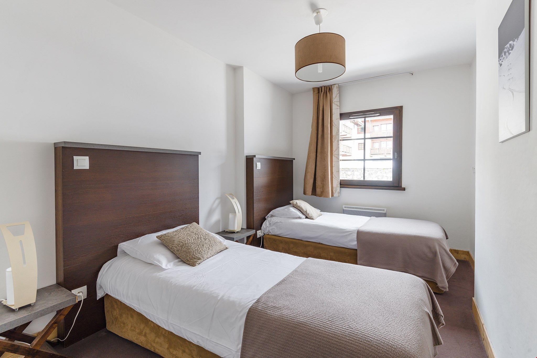 Ein Hotelzimmer mit zwei Betten, einem Schreibtisch mit Lampe, einem Fenster mit Vorhängen und einer Wandleuchte.