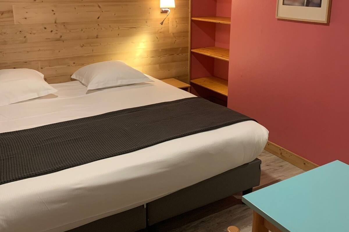 A hotel room with a bed, a bedside table, a chair, a wooden headboard, a wall-mounted shelf, and a framed picture.