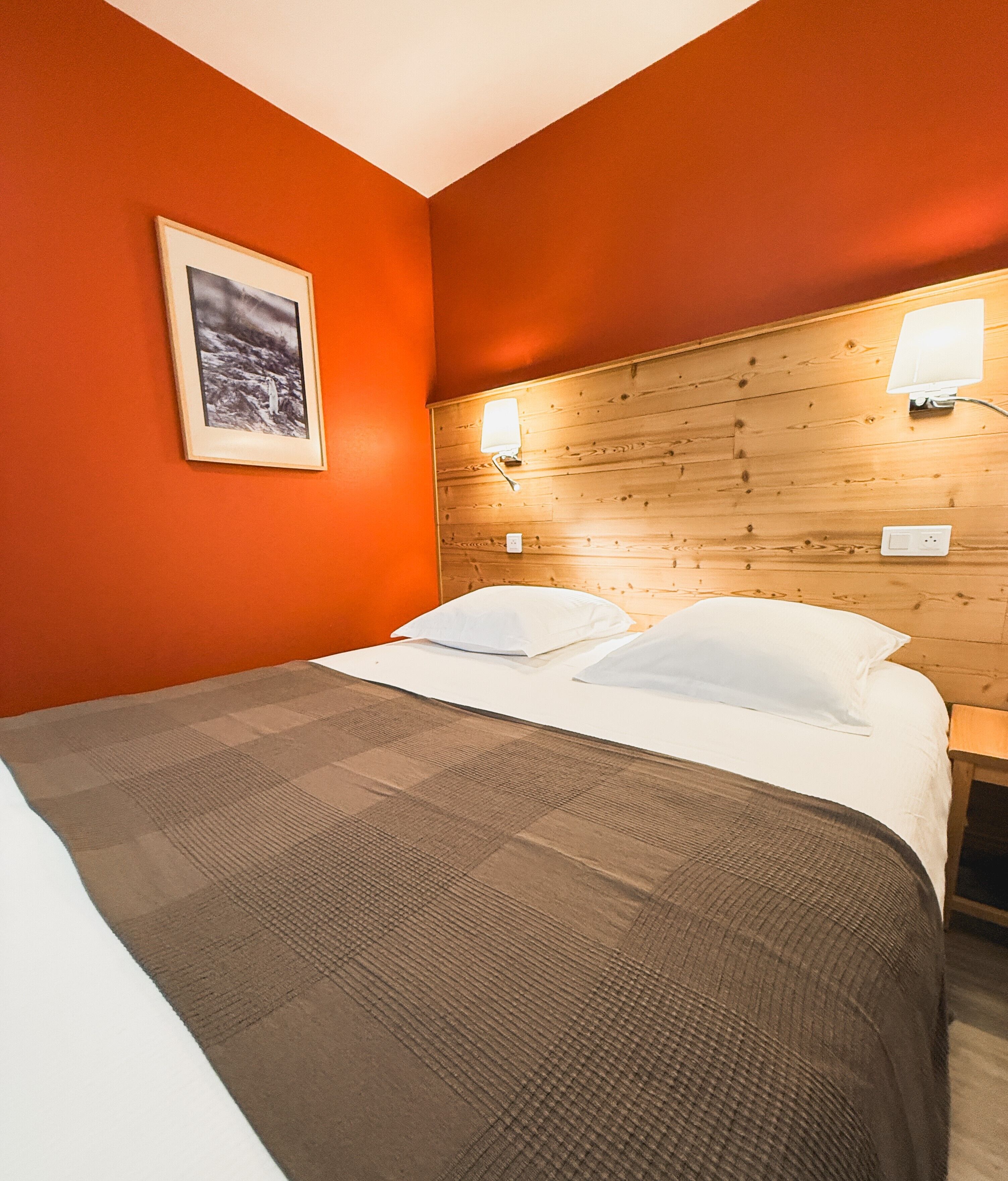 A hotel room with a wooden headboard, a bed with white linens, a brown checkered blanket, and a framed picture on the wall.