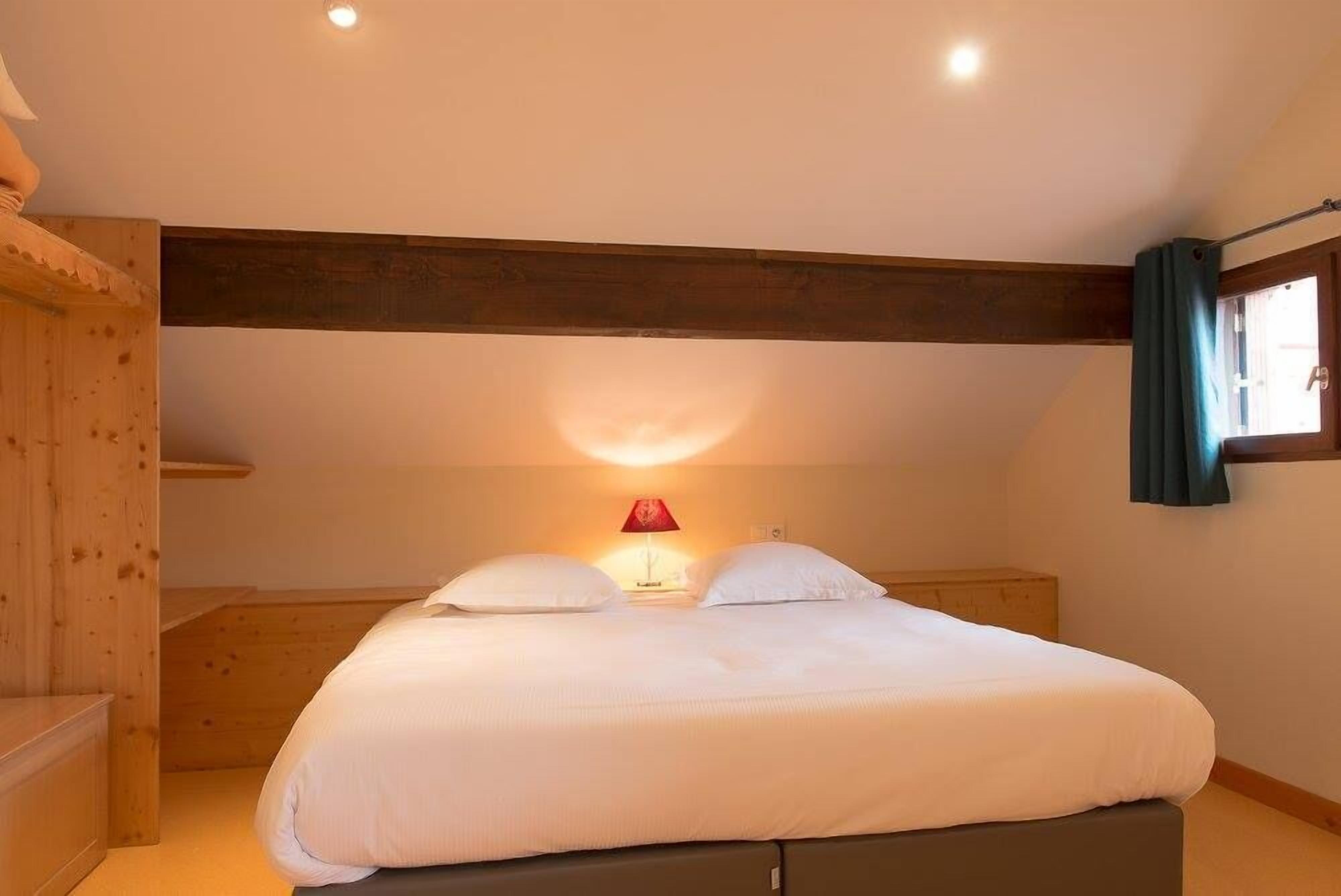 A bedroom with a large bed, wooden beams, a nightstand, a lamp, and a window with curtains.