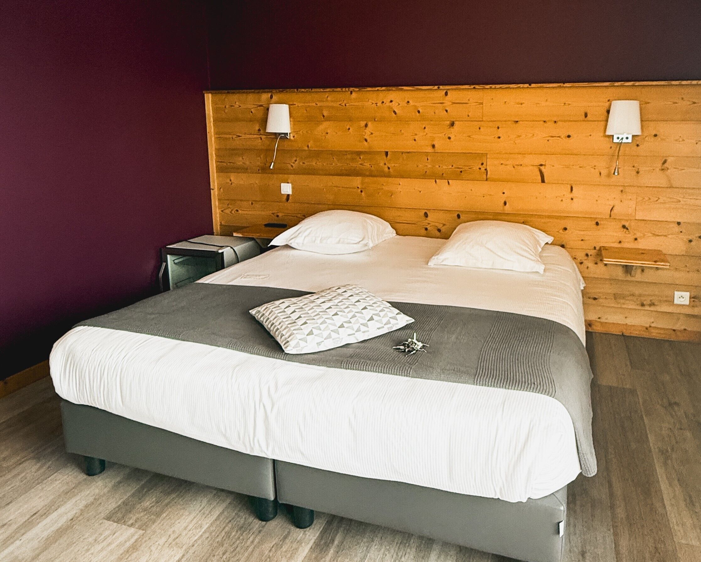 Een net opgemaakt bed met een grijze matras en witte lakens, een houten hoofdbord met wandlampen, en een klein nachtkastje met een lamp.