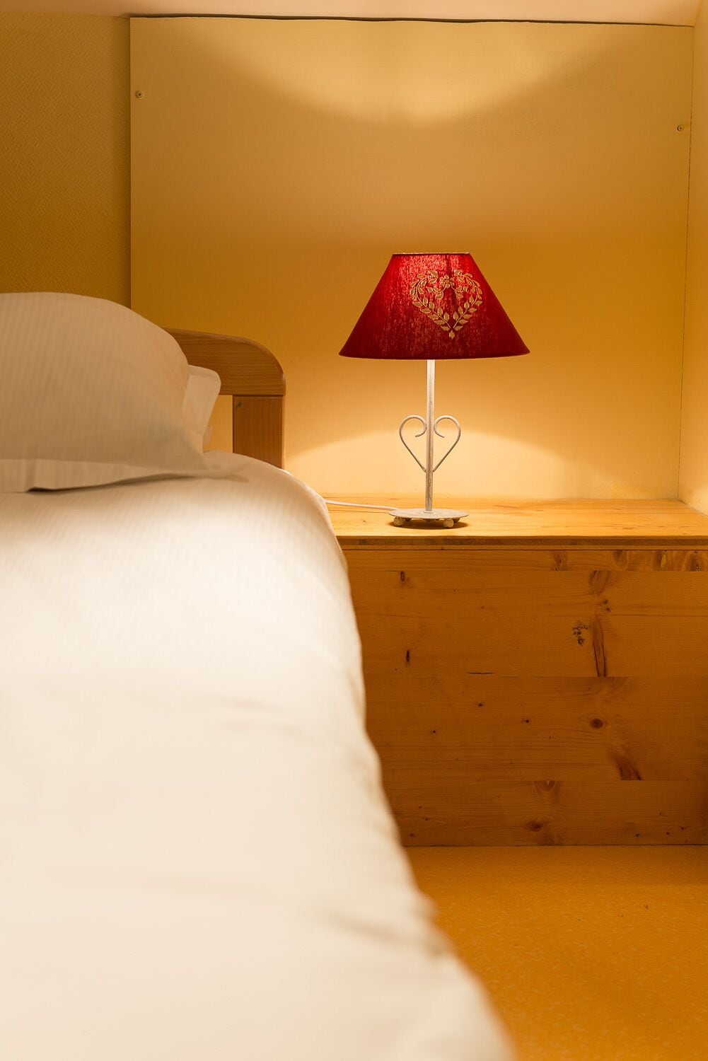 Un lit avec une literie blanche et une lampe en forme de cœur rouge posée sur une table de chevet en bois.
