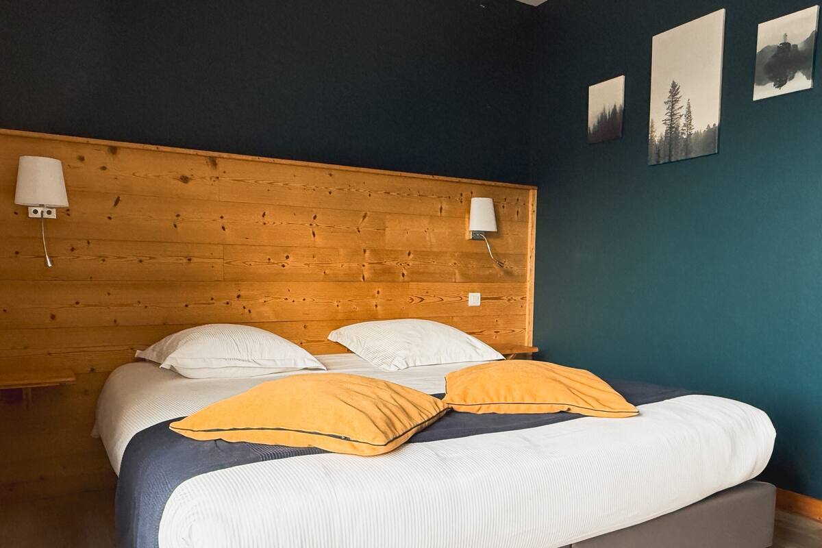A bedroom with a wooden headboard, a bed with white and yellow pillows, and a blue wall with three framed pictures.