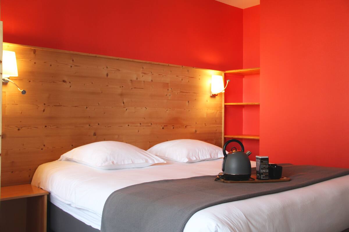 A hotel room with a wooden headboard, white bedding, a kettle, a coffee maker, and a tray with a cup and a box.