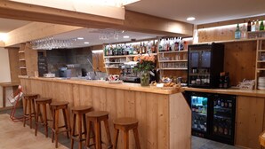 Una zona de bar con taburetes de madera, un mostrador con varias botellas, vasos y un refrigerador.