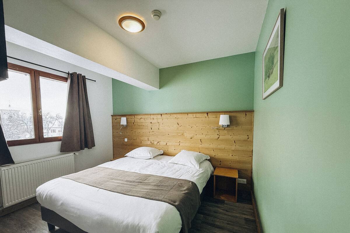 A bedroom with a wooden headboard, a window with curtains, a bedside table, and a framed picture on the wall.