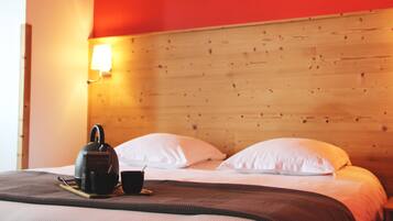 A neatly made bed with white pillows and a dark blanket, a bedside table with a kettle and a cup, and a wooden headboard with a wall-mounted light.