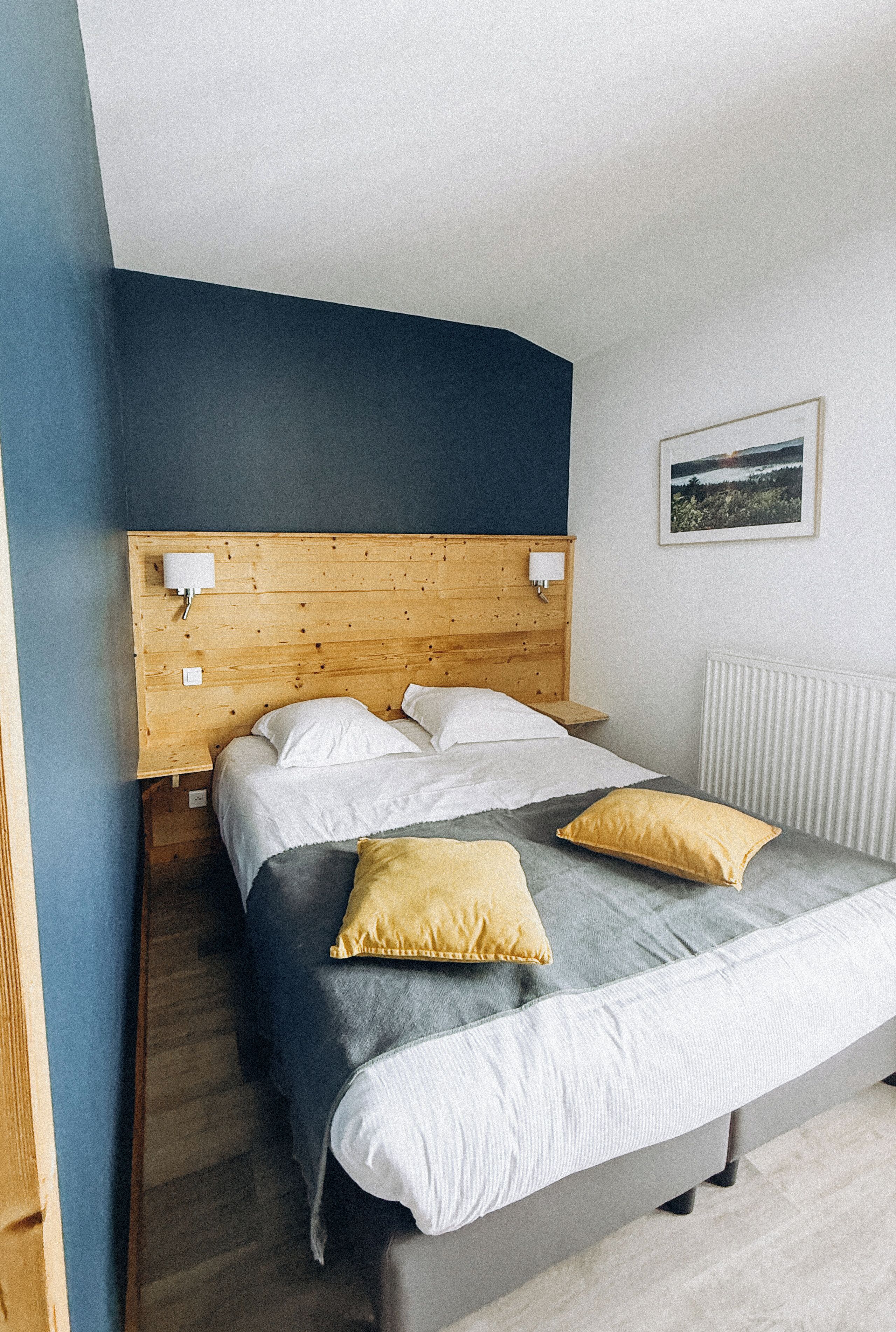 Een slaapkamer met een houten hoofdbord, een bed met wit en grijs beddengoed, twee gele kussens en een witte radiator.