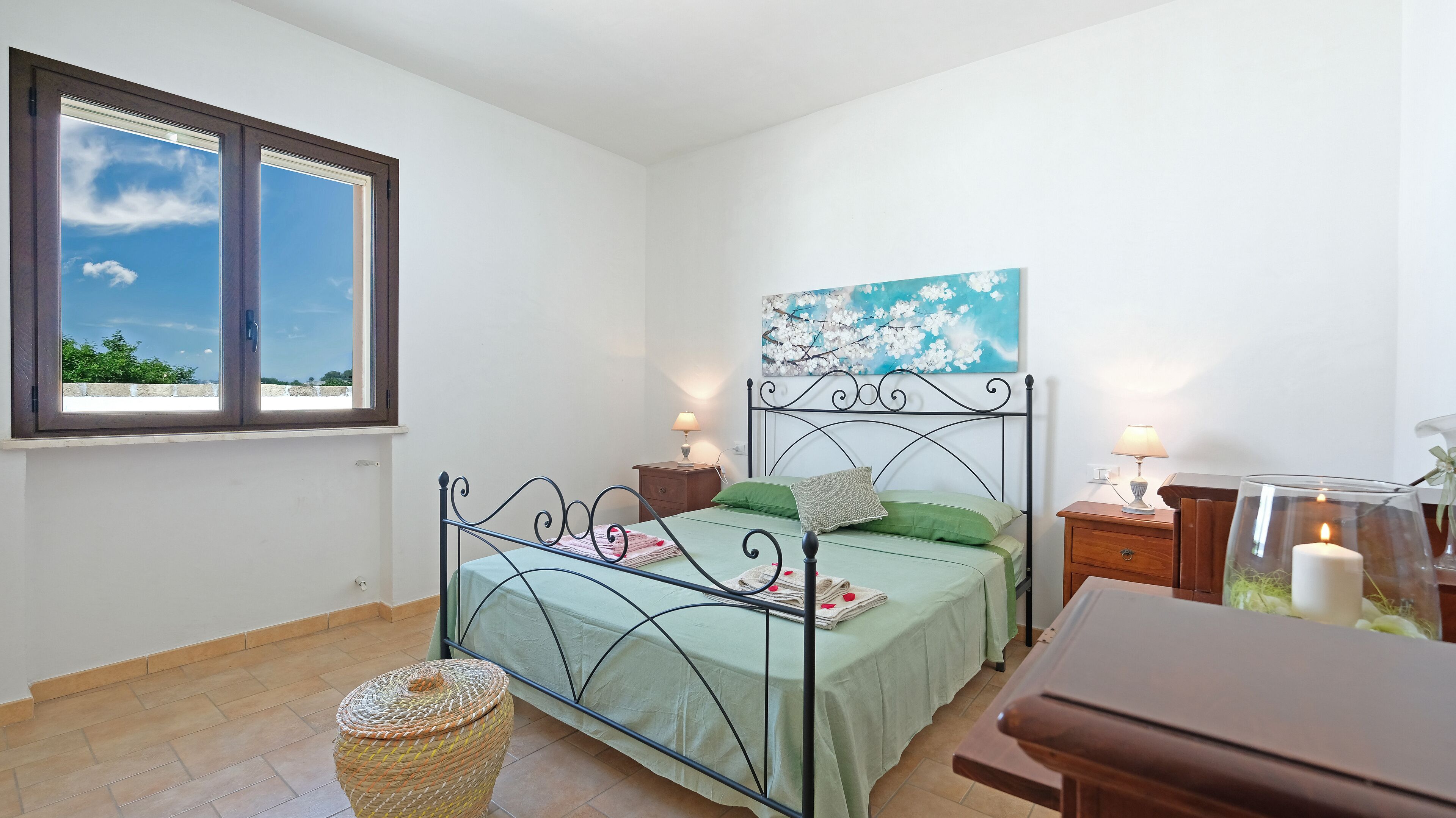 A bedroom with a bed, two bedside tables, a lamp, a painting, and a window with a view of greenery.