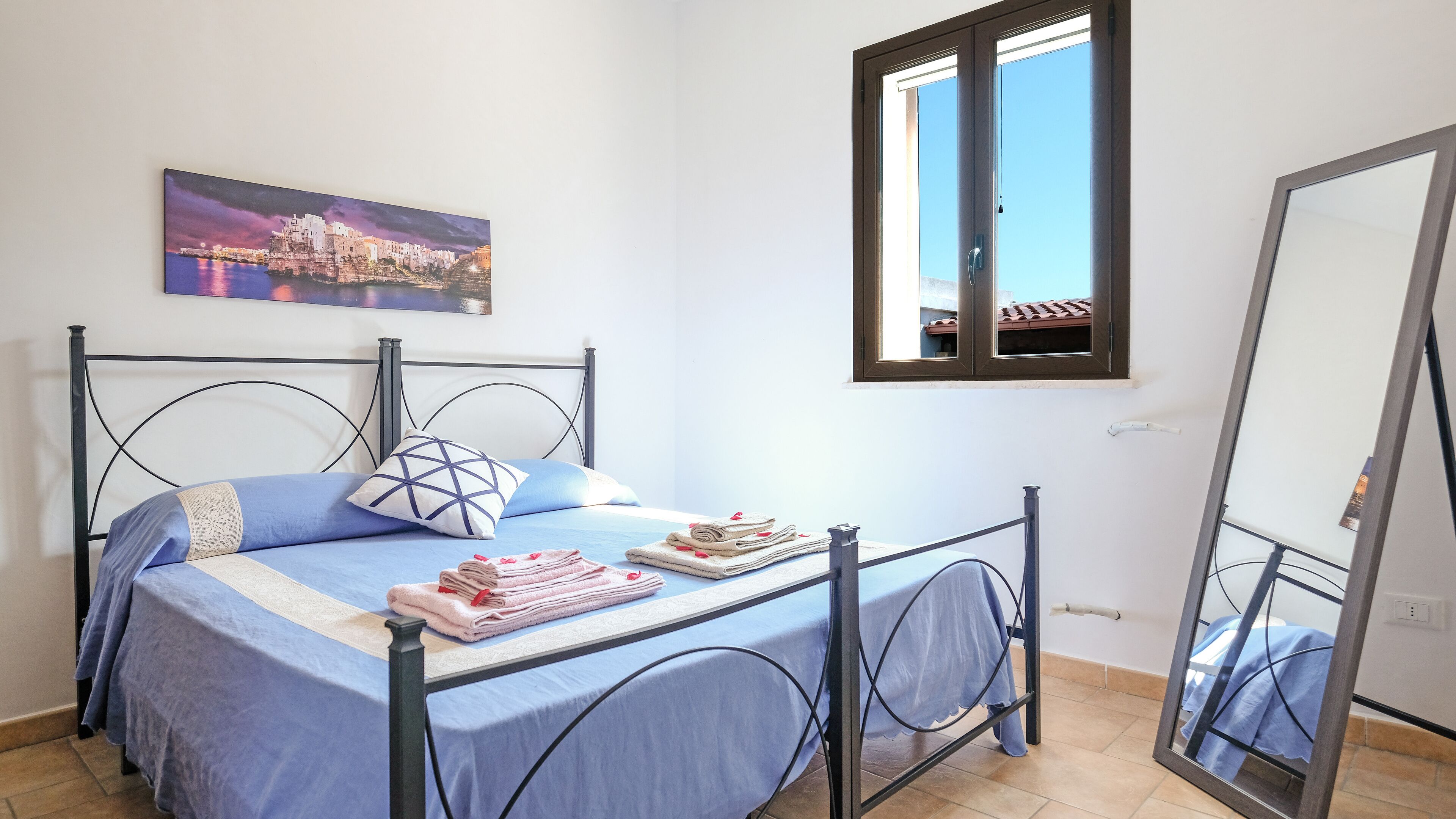 Een slaapkamer met een bed, kussens, een opgevouwen handdoek, een raam en een schilderij aan de muur.