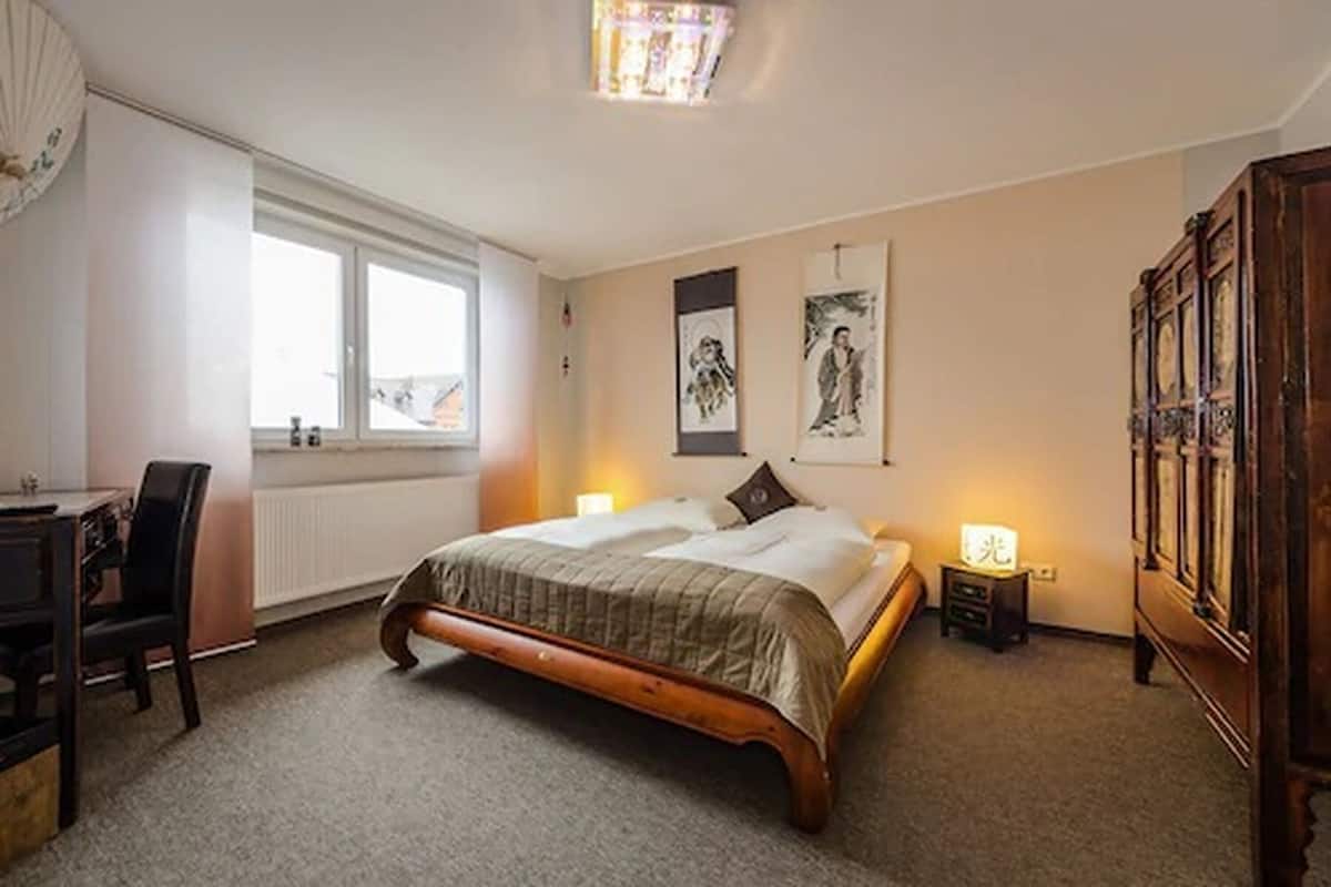 A bedroom with a wooden bed, a desk, and a chair. There is a window, a wall clock, and a cabinet.
