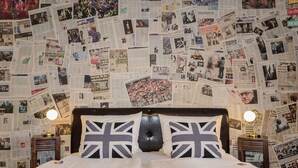 A bedroom with a bed centered in the room, flanked by two bedside tables with lamps. The walls are covered with a collage of newspaper clippings. - Hotel Rosenhof (Isenbuttel)