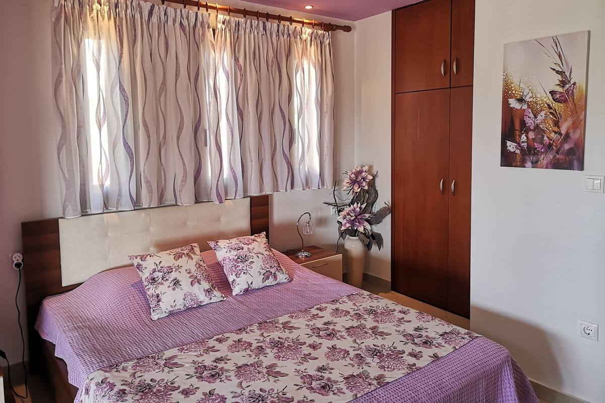 A bedroom with a bed, floral bedding, a nightstand, a vase with flowers, a wardrobe, and a painting on the wall.