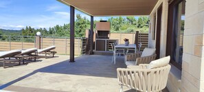 A covered outdoor patio with sun loungers, a table with chairs, and a grill.