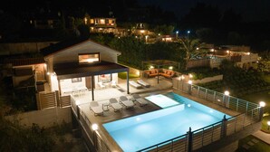 En swimmingpool med liggestole og et overdækket siddeområde, omgivet af et hegn og oplyst af lamper.