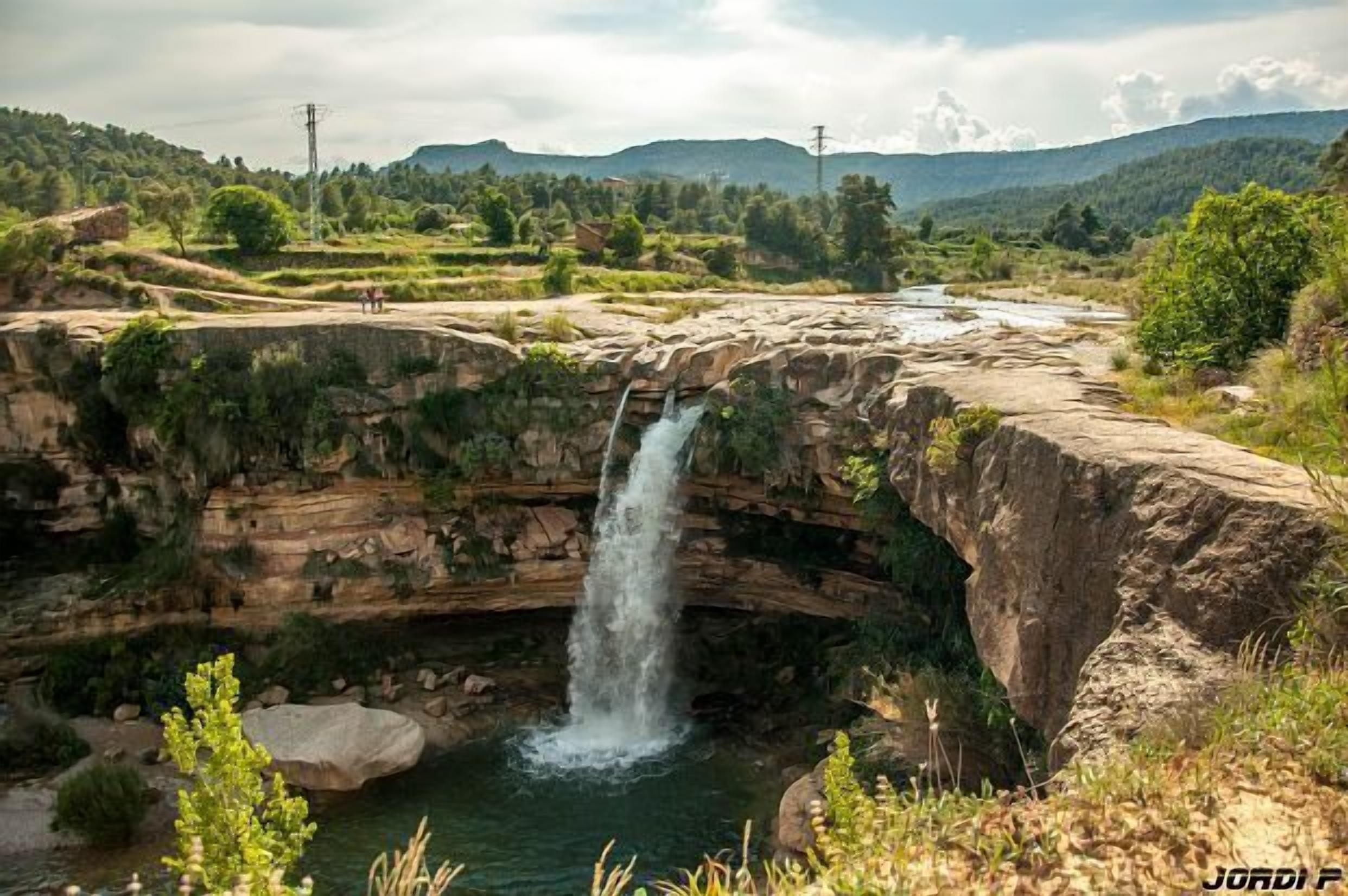 Une cascade se jetant en contrebas d’une falaise rocheuse dans une rivière, le tout entouré d’une végétation luxuriante et de collines lointaines.